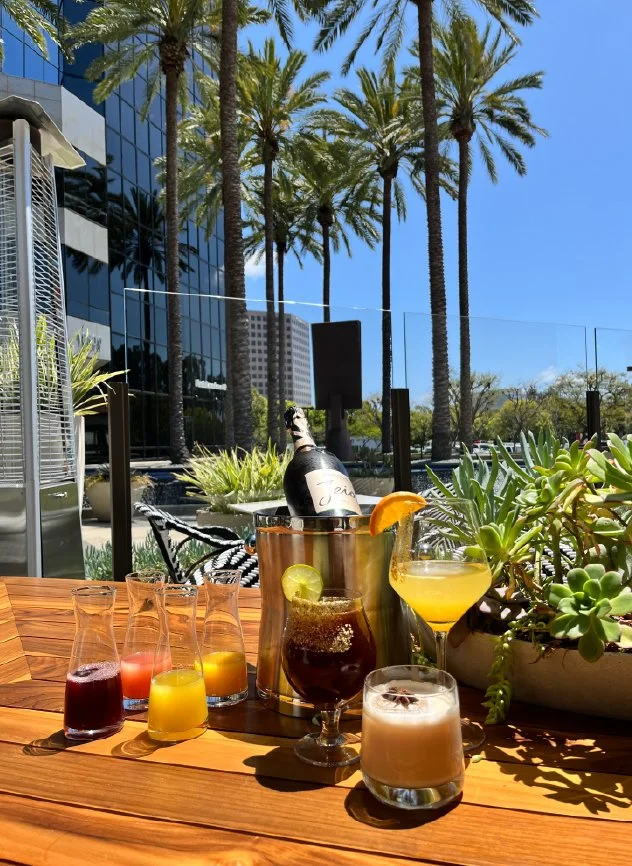 Assorted colorful drinks on a wooden table outdoors, with palm trees and a building in the background on a sunny day.