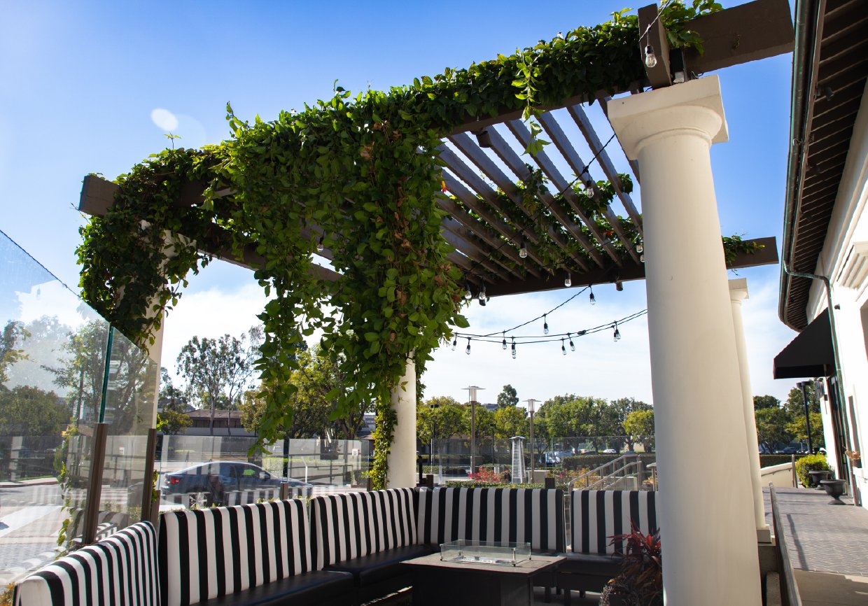 Outdoor seating area with black and white striped cushioned benches, a glass-topped table, white columns, a wooden pergola with vines, string lights, and a view of trees and parked cars under a blue sky.