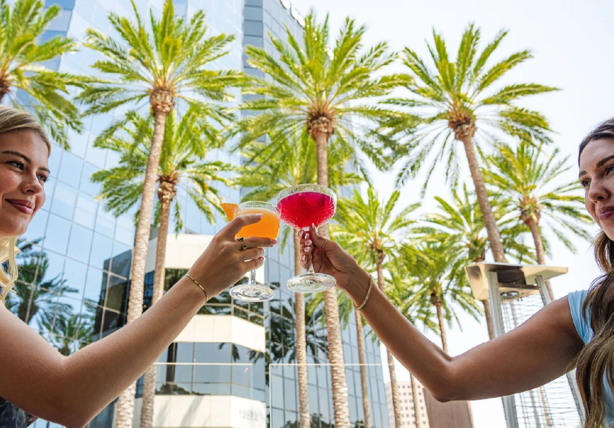 Two women clinking colorful cocktails outdoors with palm trees and a glass building in the background.