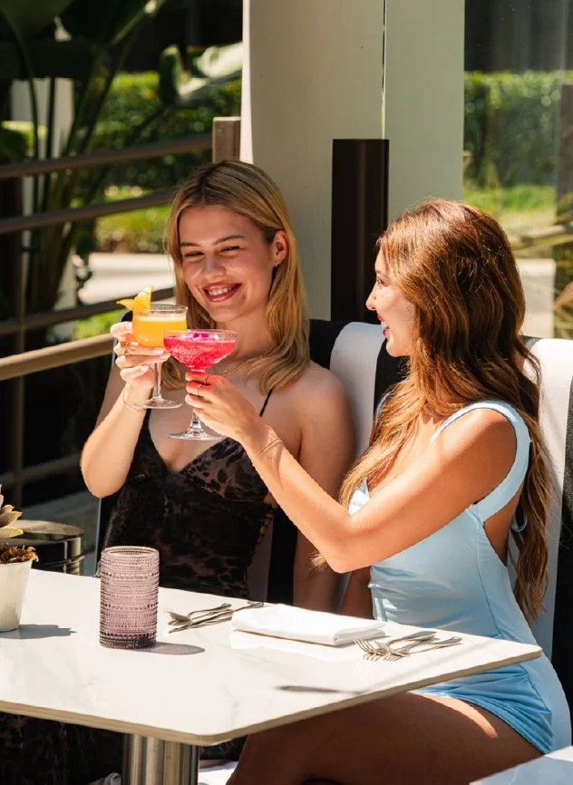 Two women celebrating with cocktails at a restaurant table outdoors, smiling and toasting each other's drinks.