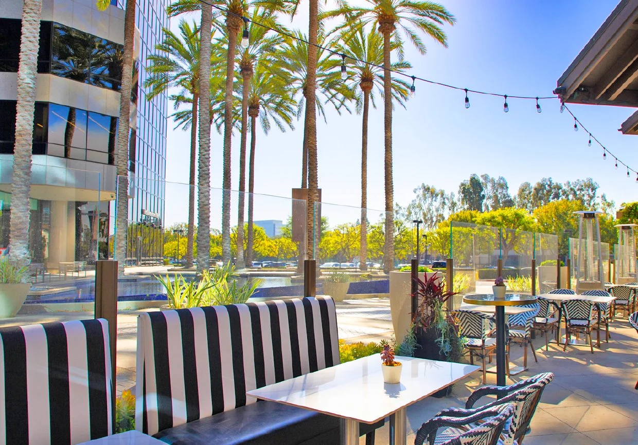 Outdoor patio seating area at a restaurant with striped chairs, small tables with plants, and glass partitions. Tall palm trees and a modern glass building are visible in the background on a sunny day.