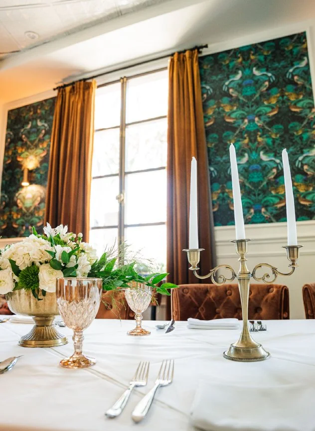 Elegant dining table set for a formal event with a floral centerpiece, gold candlestick holder with three white candles, pink glassware, silverware, and napkins, with large window with gold curtains and decorative wallpaper in the background.