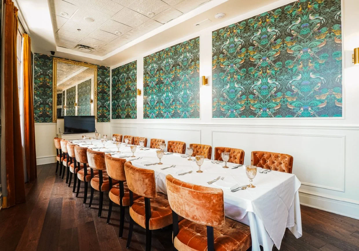 A long dining table set with white tablecloths, glassware, and silverware, surrounded by orange velvet chairs, in a room with vibrant patterned wallpaper and gold wall sconces.