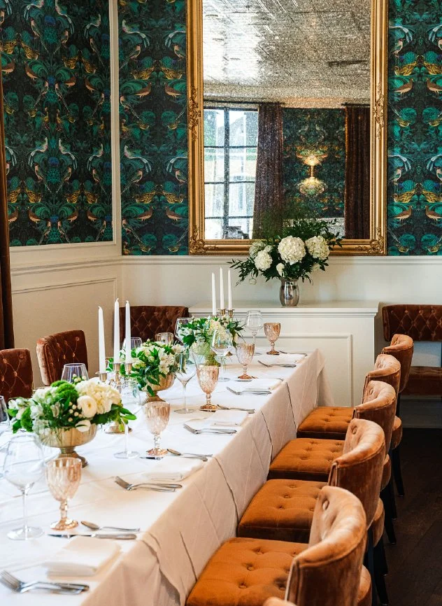 Elegant dining table set for a formal event with white tablecloth, floral centerpieces, candles, glassware, and gold-rimmed glasses in a decorated room with patterned teal wallpaper and a large mirror