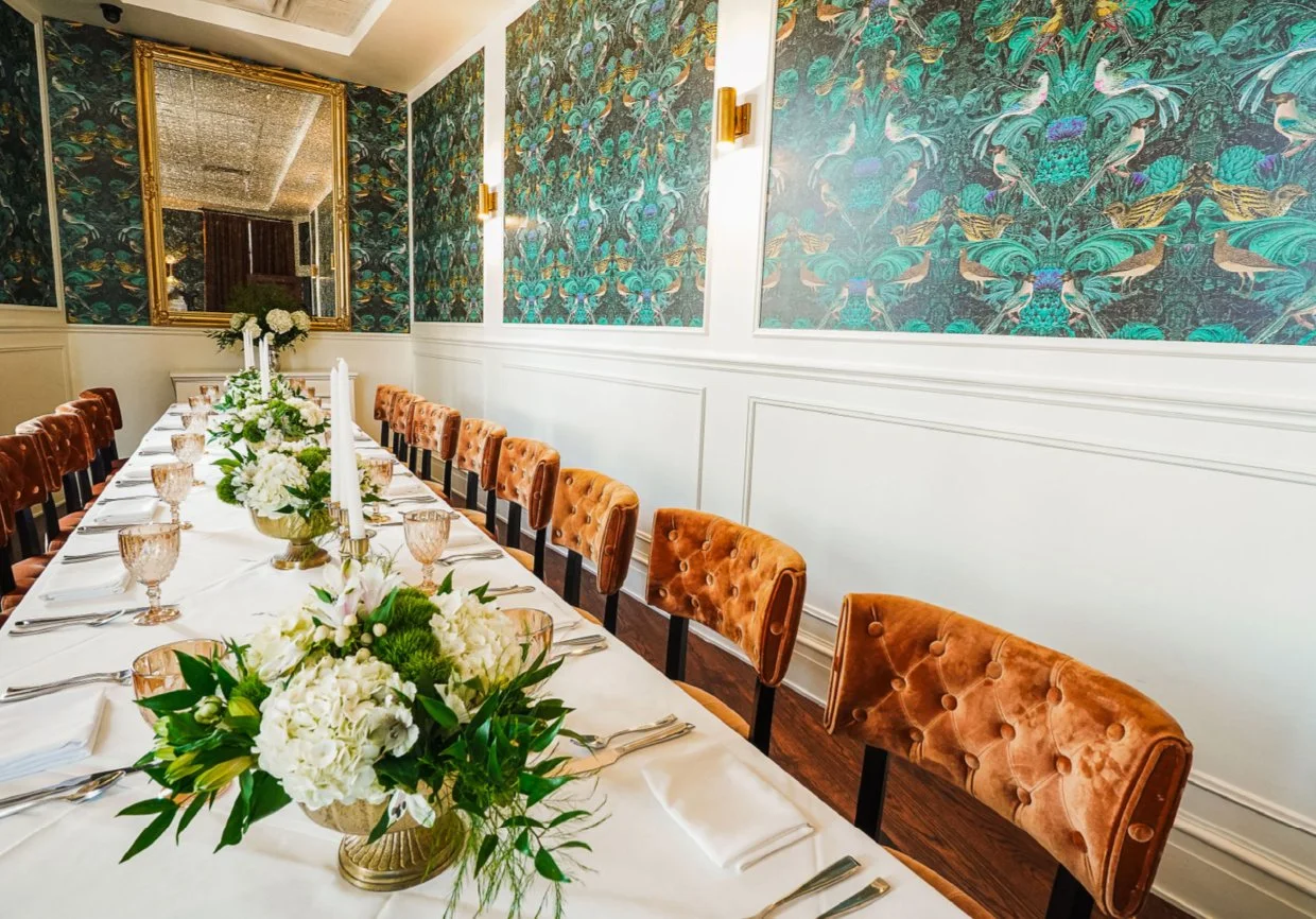 A long dining table set for a formal event with white tablecloths, floral centerpieces, pink glasses, and silverware. Orange velvet chairs surround the table. The room has ornate floral wallpaper, white wainscoting, and a large mirror on the wall.