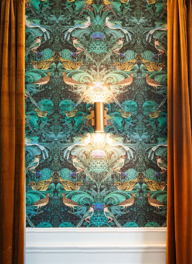 Decorative wall with tropical bird and plant pattern, illuminated by a wall sconce, framed by mustard-colored curtains.