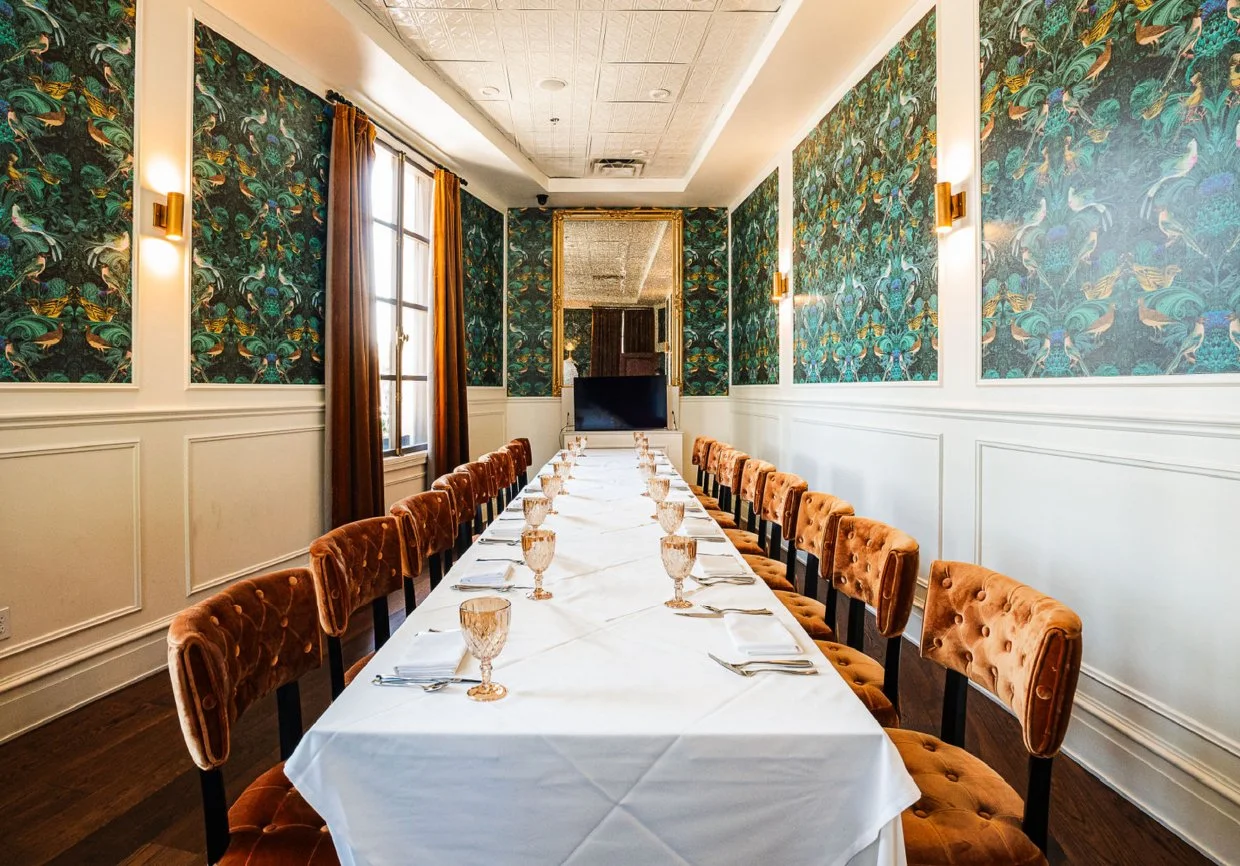 A long rectangular dining table set with white tablecloth, glassware, and silverware in a decorated dining room with patterned wallpaper, velvet chairs, and a large mirror on the wall.