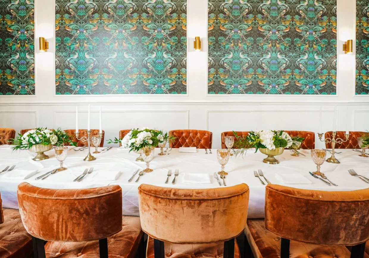 A formal dining room with a long table set with white tablecloth, floral centerpieces, candles, and glassware, surrounded by upholstered chairs. The wall features a large, intricate decorative wallpaper design with gold accents and wall sconces.