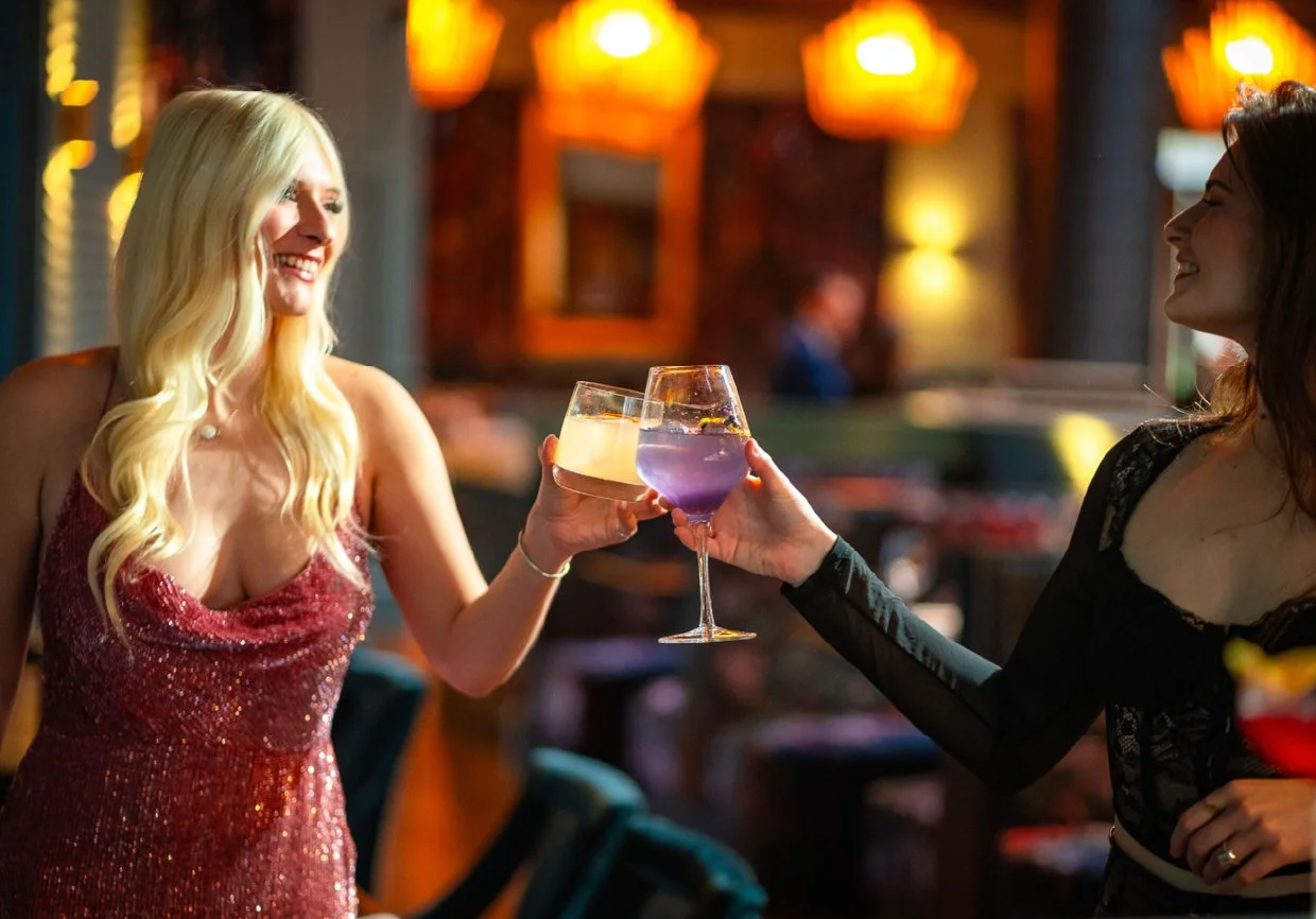 Two women clinking glasses of colorful drinks in a dimly lit bar or restaurant.
