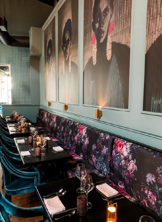A restaurant interior with a long table set for dining, featuring wine glasses, candles, and napkins. The wall has large artistic portraits of women with dark hair and pink earrings, and there are velvet chairs with floral patterns and others in teal