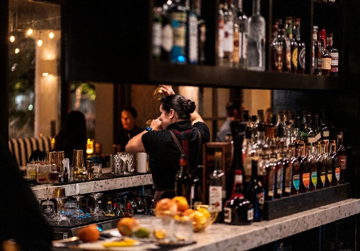 A bartender mixing drinks behind a bar with various bottles of alcohol, with patrons in the background and fruit on the counter.