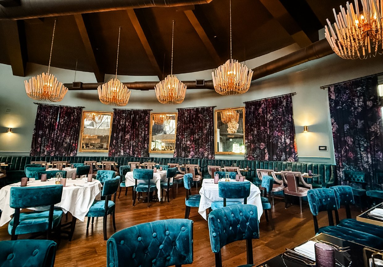 Interior of a restaurant with blue velvet chairs, tables with white tablecloths, dark floral curtains, and chandeliers hanging from a wooden ceiling.