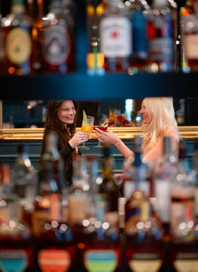Two women in a bar toasting with drinks, blurred bottles in the foreground, smiling and enjoying their time.