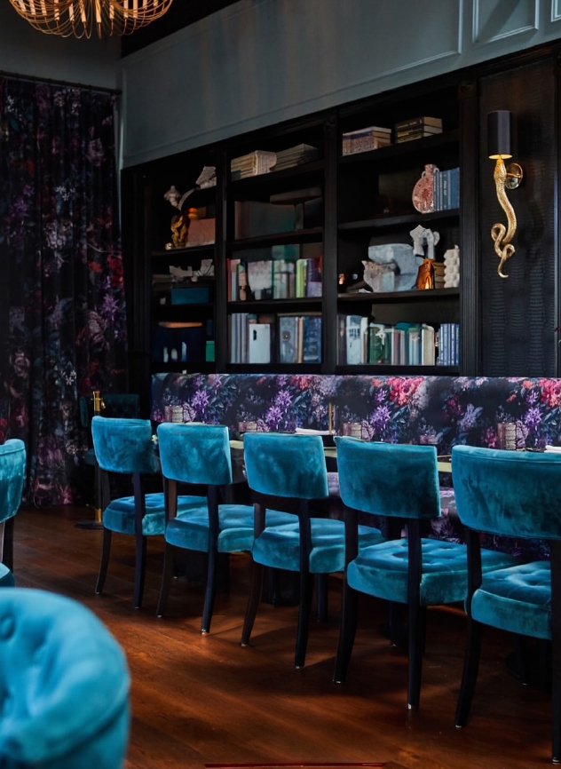 Interior of a stylish room with a dark wooden bookshelf filled with books and decorative objects, dark floral curtain, and a row of plush teal velvet chairs along a floral upholstered bench, with wooden flooring.
