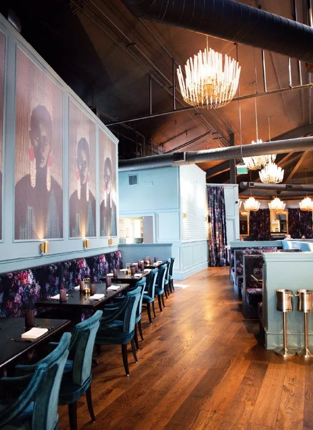 Interior of a restaurant with blue walls, a floral patterned banquette, dark wood tables set with placemats and glasses, teal upholstered chairs, and elegant chandeliers hanging from a dark ceiling.