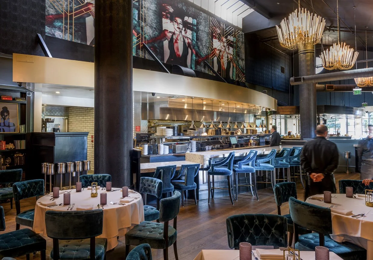 Interior of a modern restaurant with a bar, round tables with chairs, and chandeliers. There are paintings on the wall and open kitchen area with staff preparing food.