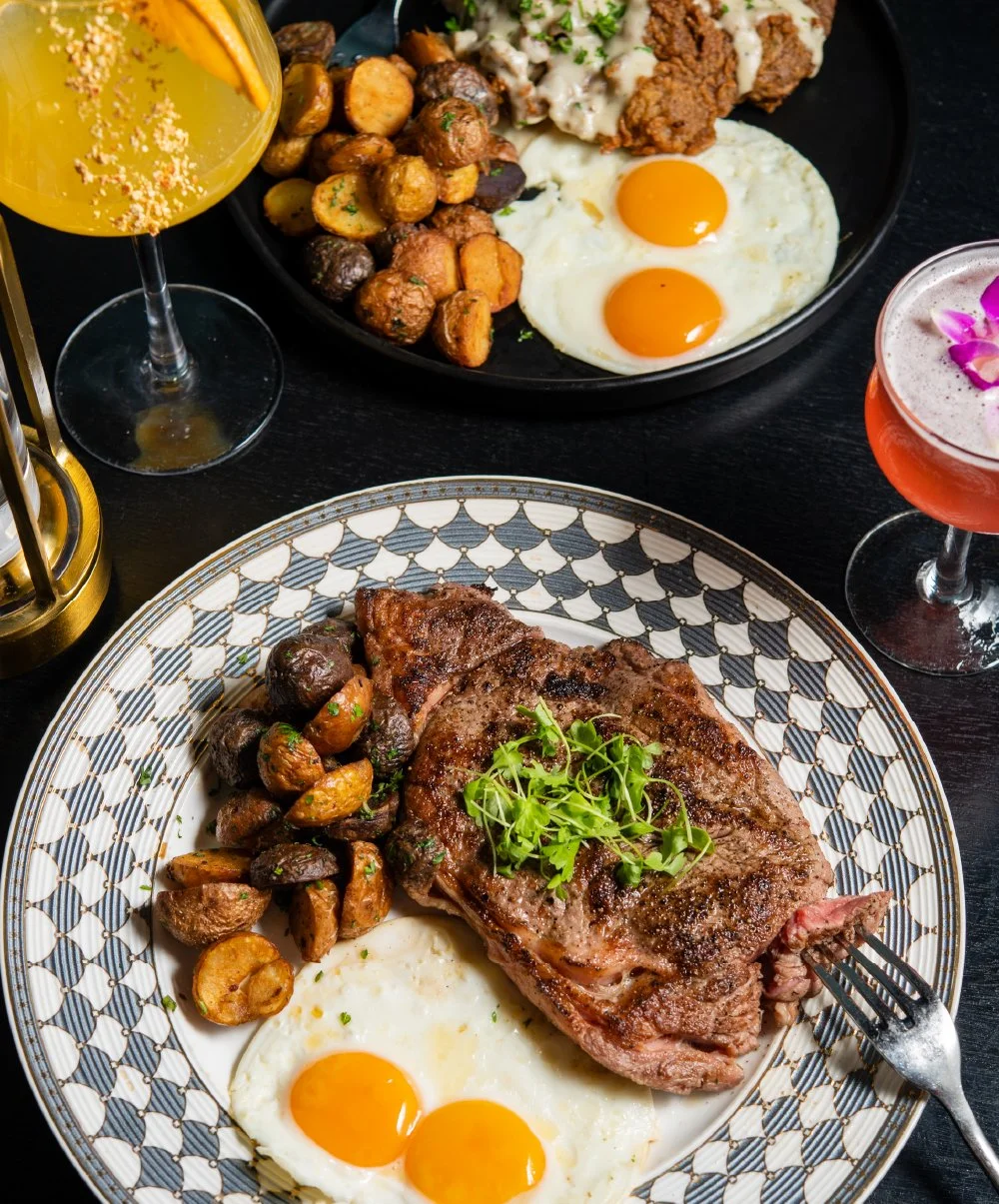 A plate with a grilled steak topped with microgreens, fried eggs with runny yolks, and roasted potatoes. In the background, a black plate with fried chicken, sautéed vegetables, and scrambled eggs. Two colorful cocktails with flowers are also on the table.