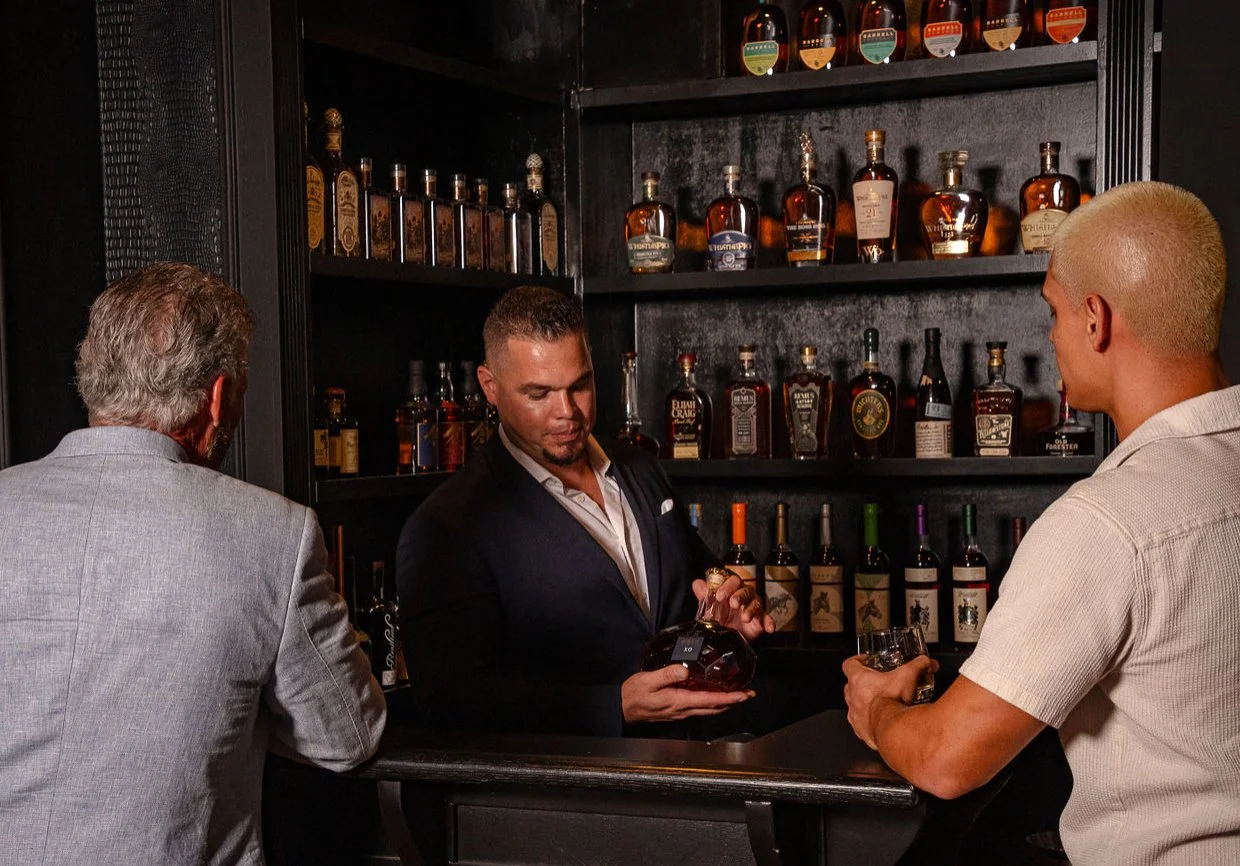 Three men at a bar, one bartender pouring whiskey from a bottle into a glass, with shelves of liquor bottles behind them.