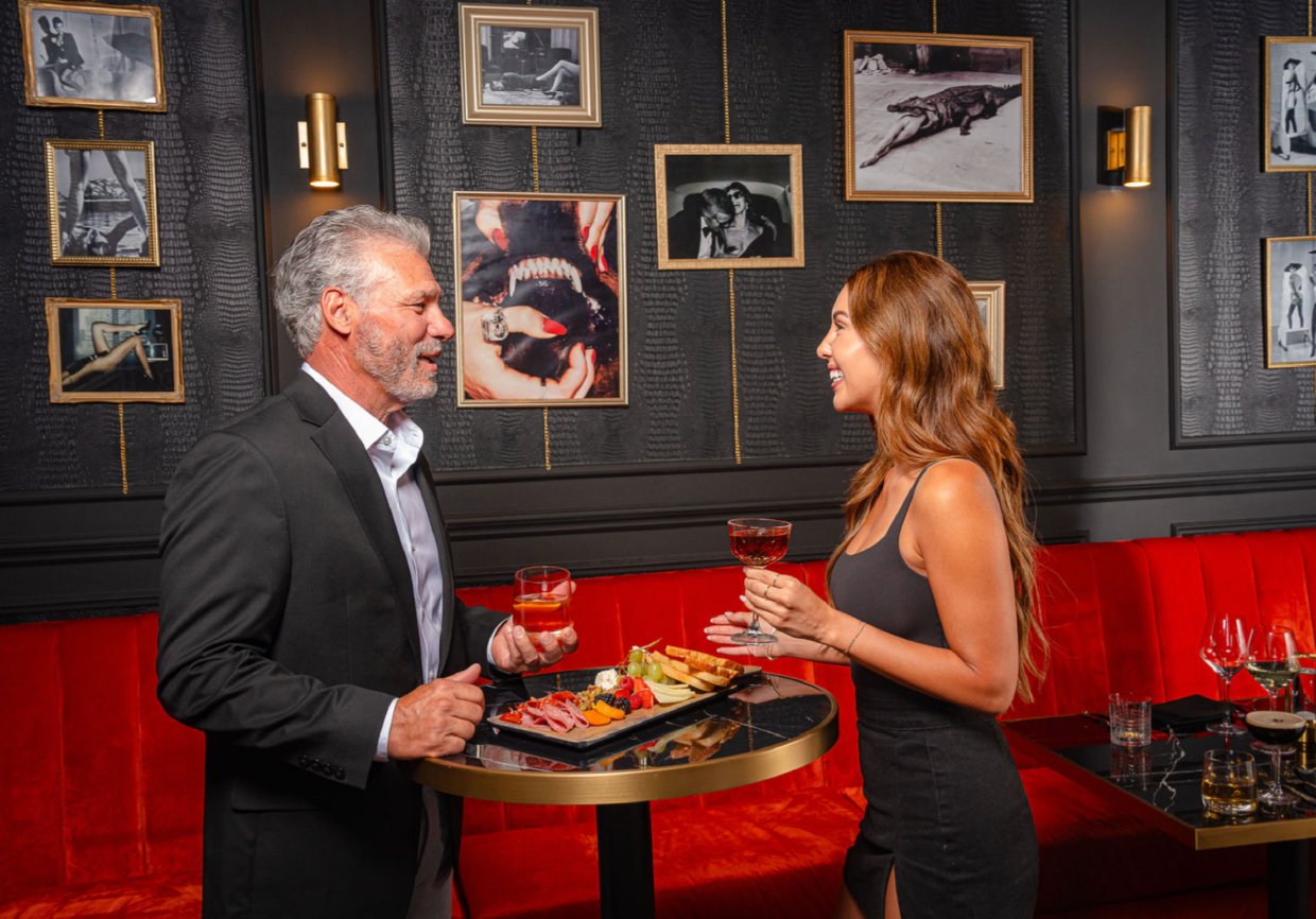 A man and woman chat and smile in a restaurant, with the man holding a drink and a tray of appetizers, and the woman holding a wine glass, surrounded by framed photographs on a dark wall.