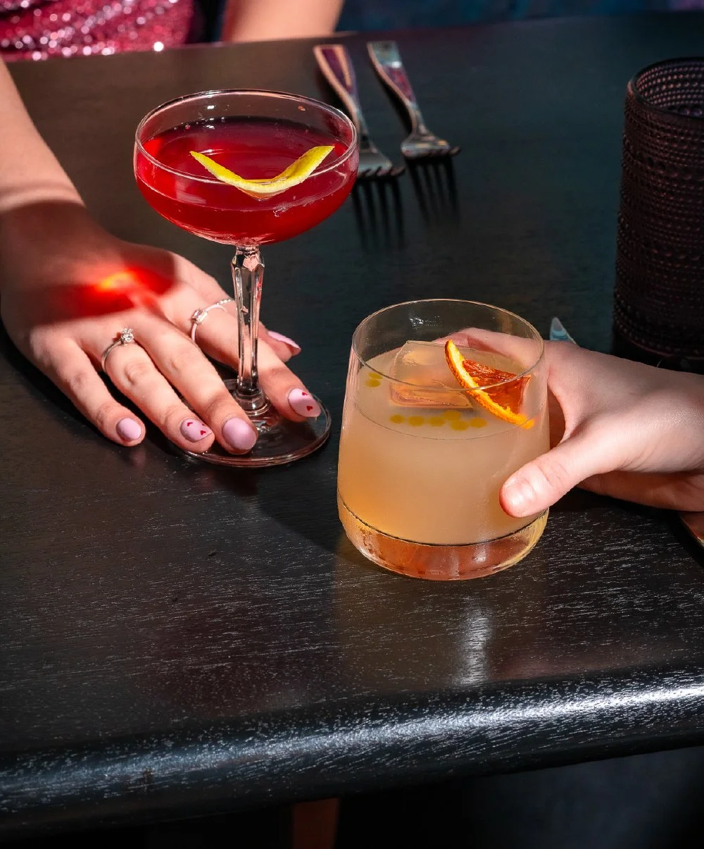 Two people clinking cocktails at a dark-colored table, one with a pink-colored drink and the other with a yellow-colored drink, garnished with citrus slices.