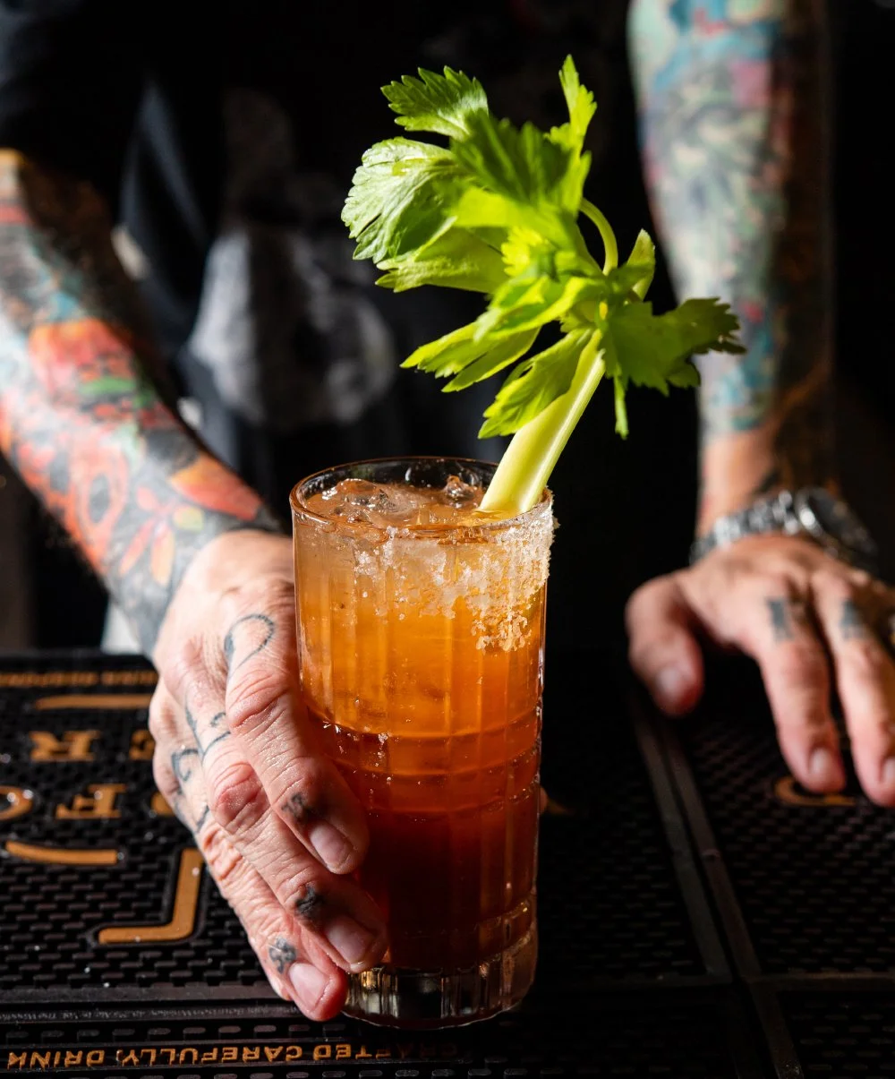 A person with tattoos on their arms holding a tall cocktail glass with a drink garnished with a stalk of celery.