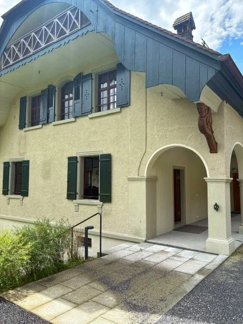 Exterior of a yellow house with blue trim, green shutters, and an arched entrance, featuring a wooden decorative element near the roof and a small pathway leading to the door.