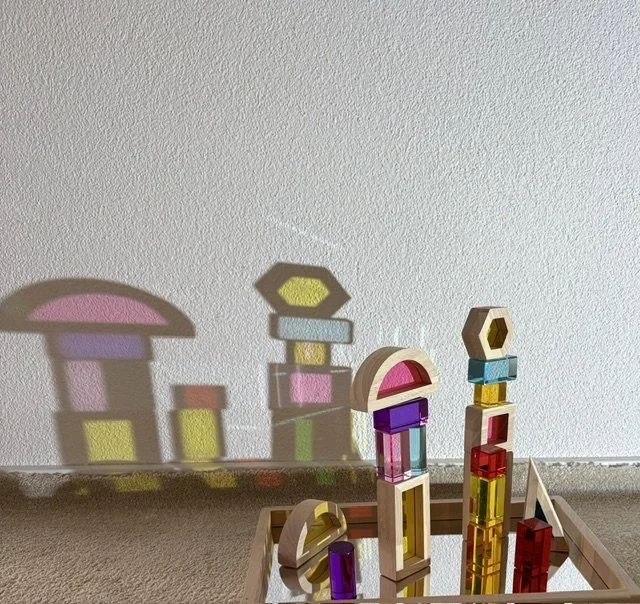Colorful wooden building blocks arranged on a surface, casting playful shadows on a white wall.
