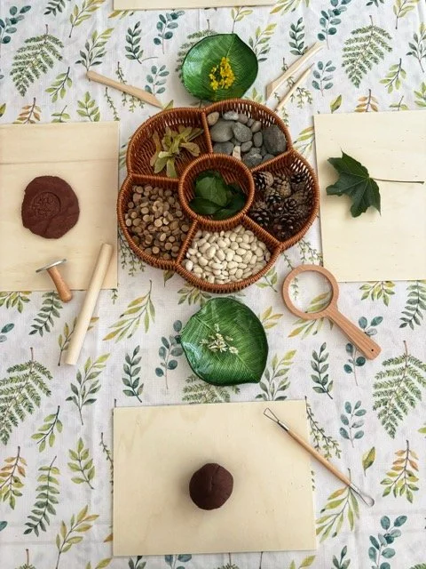 A flat lay of nature-inspired crafting supplies on a leaf-patterned tablecloth, including a container with small stones, leaves, pinecones, and white beans, along with notebooks, pens, and decorating tools.