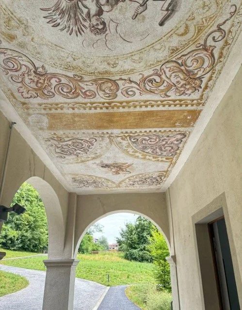 Ceiling with ornate, classical-style fresco artwork on a covered outdoor porch with arches and green landscape in the background.