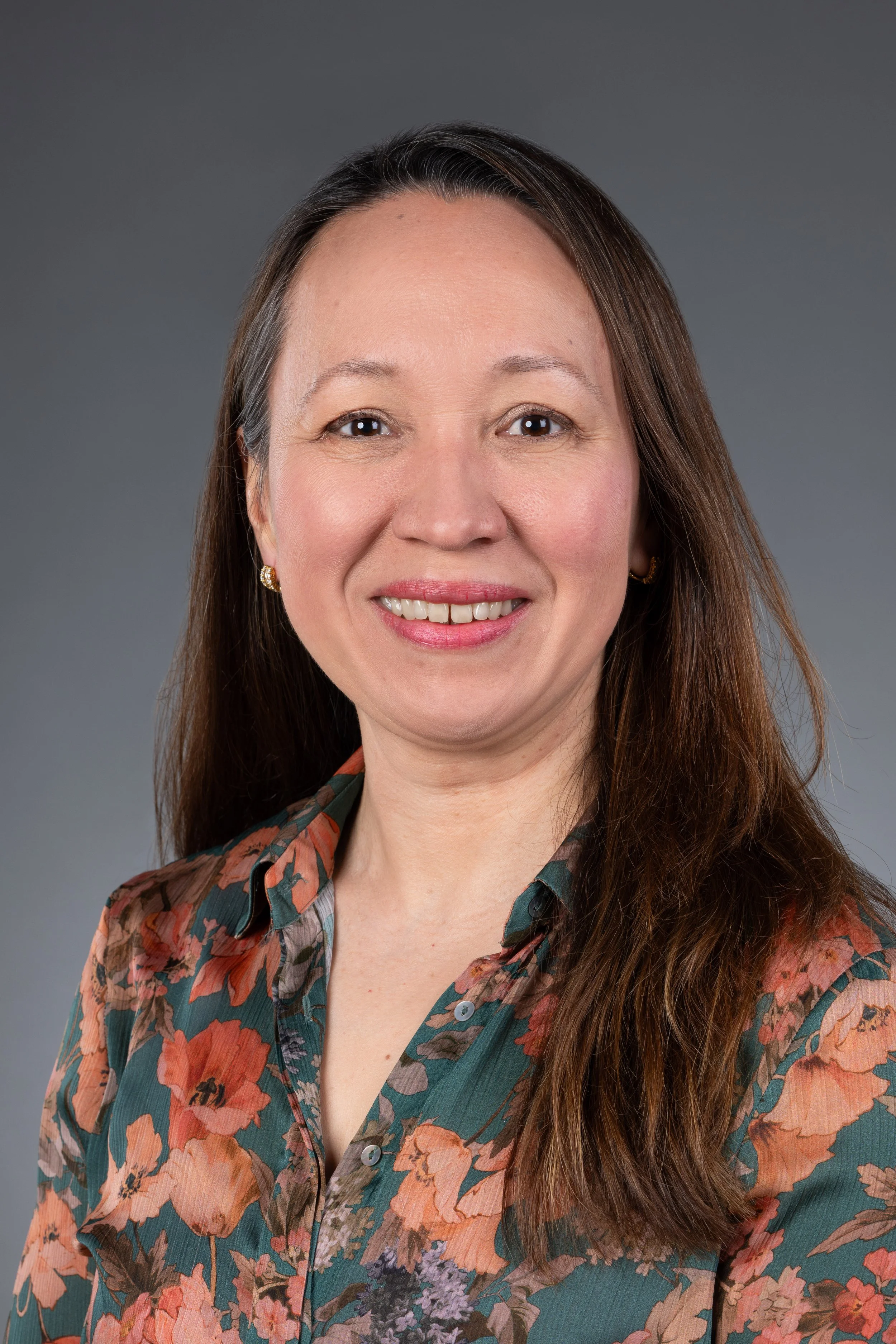 A woman with long brown hair smiling, wearing a floral patterned shirt against a neutral gray background.
