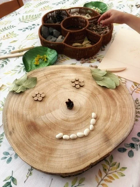A wooden tree slice with white and brown beans arranged to form a smiley face, with three leaves on each side as eyebrows, on a table with botanical patterned cloth. In the background, a hand reaches toward a divided basket of stones and small objects, and a green leaf with yellow flowers is nearby.