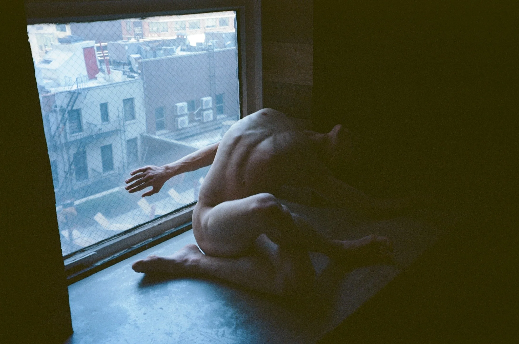 A person sitting on the floor by a window, reaching out with their right arm, with the cityscape outside visible through the window, featuring buildings and a fire escape.