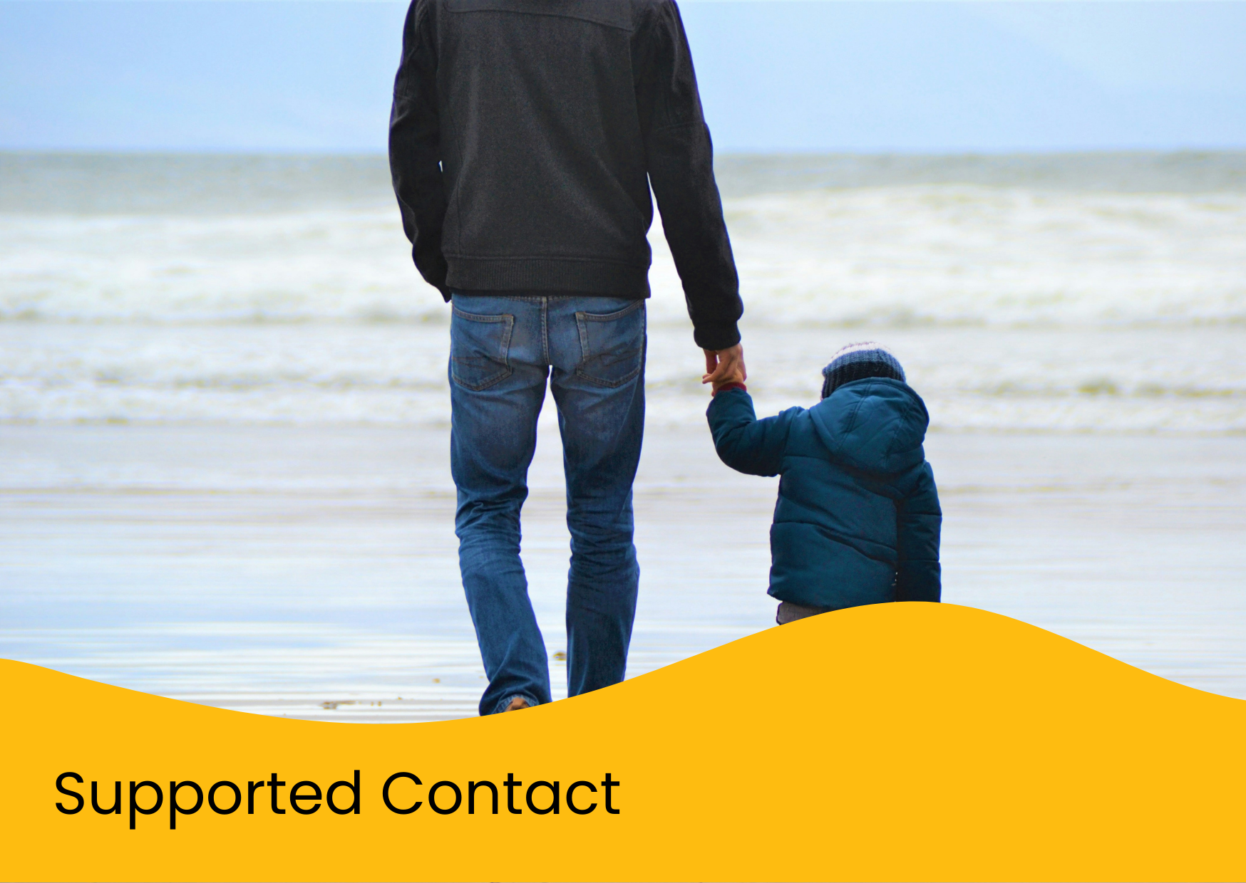 A man and a young child walking hand in hand on the beach, with the ocean in the background.