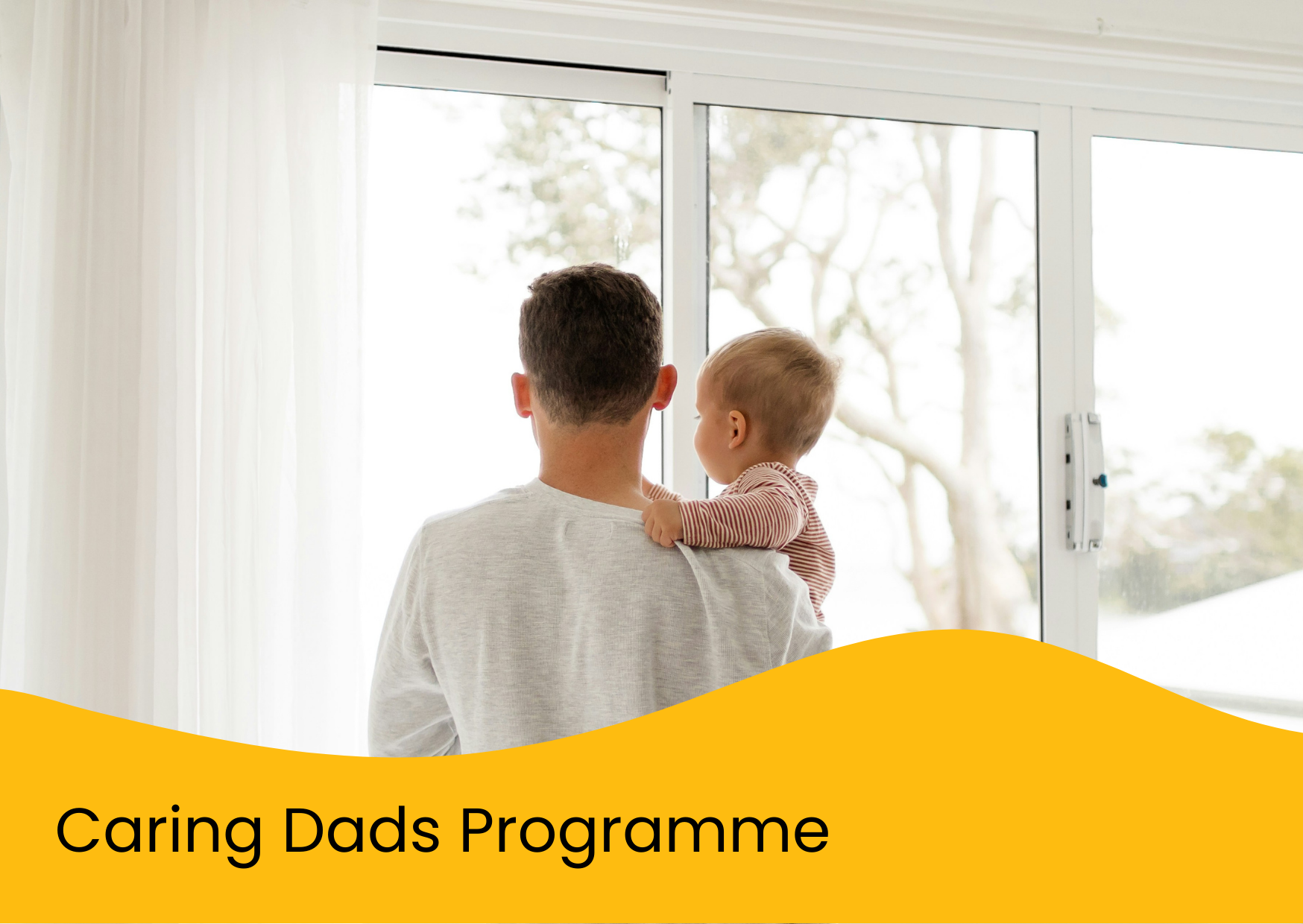 A father holding his young child while looking out a large window with white curtains, with trees visible outside.