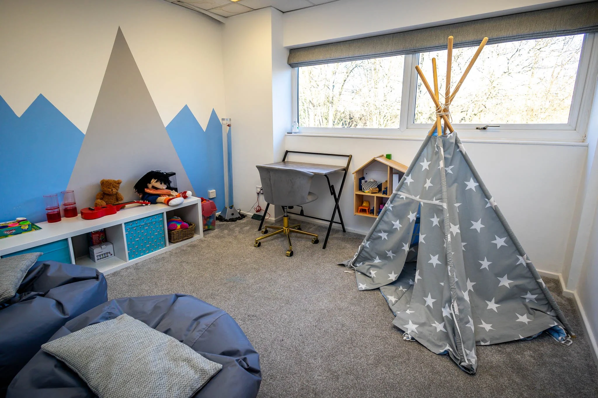 A children's playroom with a white wall painted with blue mountain peaks, a white shelf with toys and red cups, stuffed animals, a desk with a chair, a small wooden house, a teepee tent with a star pattern, a window with a view of trees outside, and a gray bean bag chair with pillows on the carpeted floor.