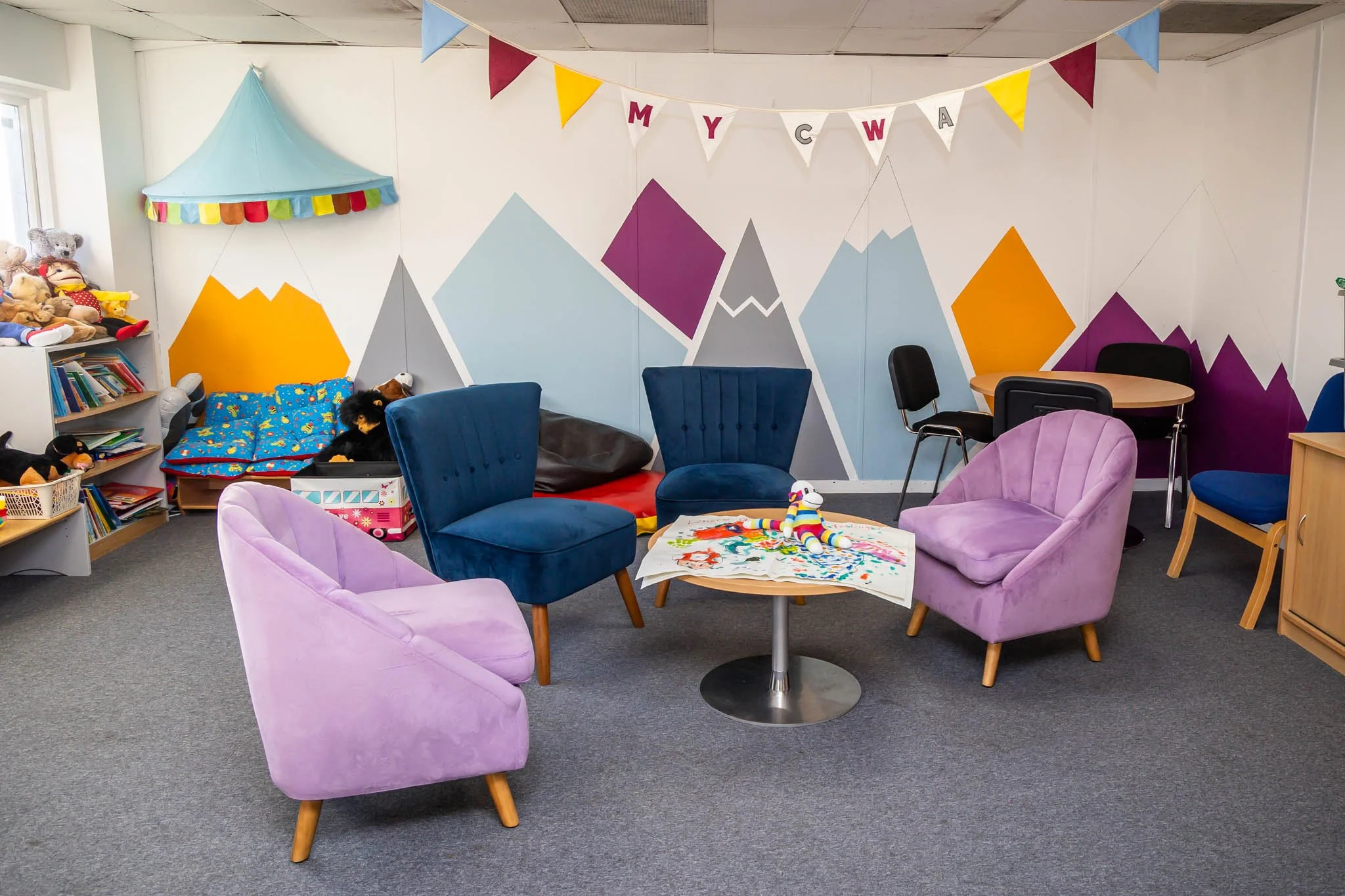 A cozy indoor playroom decorated for a celebration with colorful bunting banners and a mural of mountain peaks painted on the white wall. The room contains purple and blue armchairs, a small table with a children's drawing and a rainbow-striped stuffed animal, a bookshelf filled with toys and books, and a corner with a tent and plush toys, all on a gray carpeted floor.