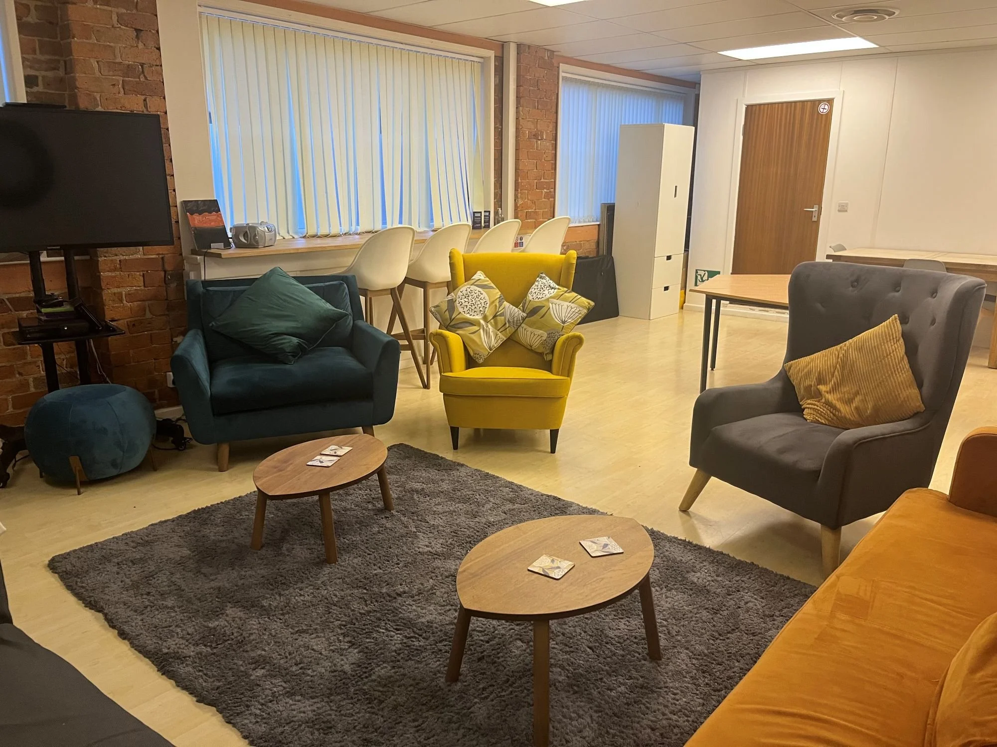 A cozy lounge area with several armchairs, a small wooden table, and a dark rug. There are large windows with vertical blinds, a brick wall, a television on a stand, and various decorative pillows.