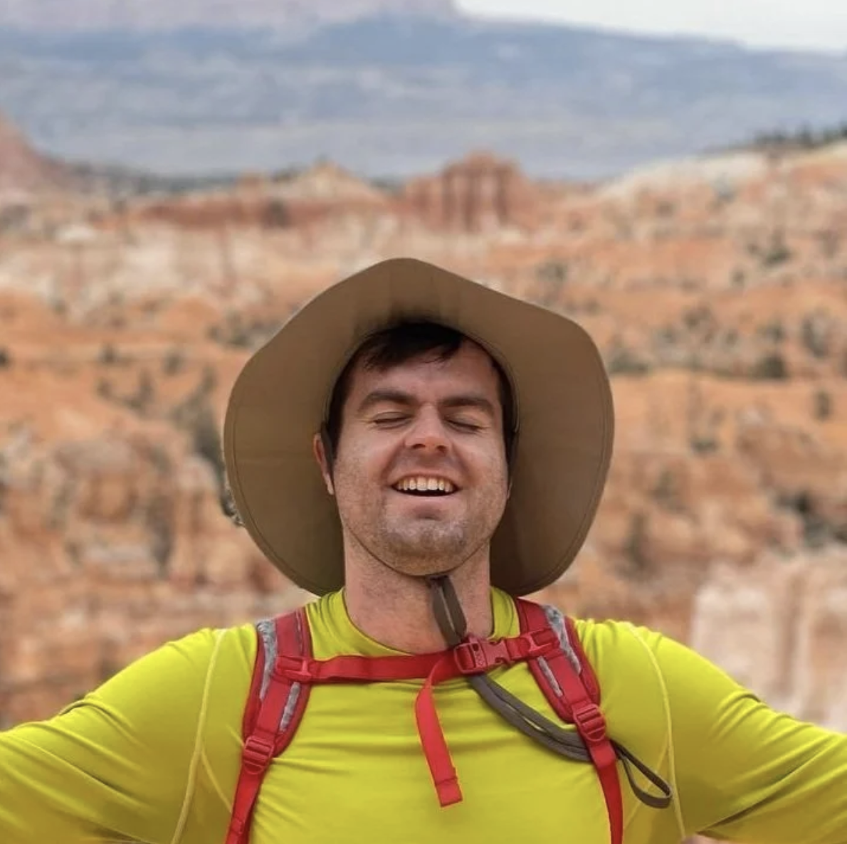 A man with a wide-brimmed hat smiling in front of desert canyon landscapes, wearing a yellow shirt and red backpack.