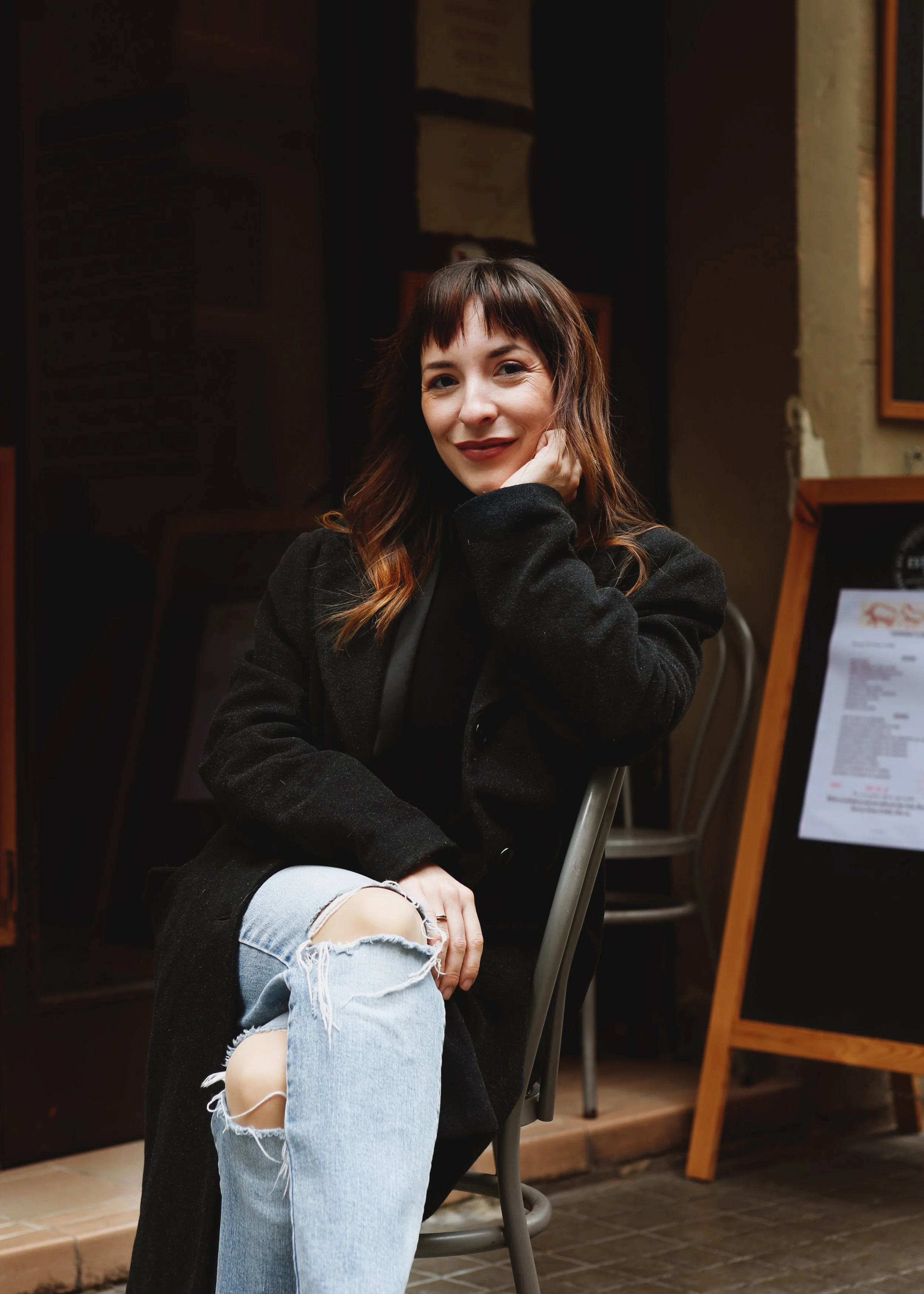 A young woman with shoulder-length brown hair sitting in a chair, smiling slightly, resting her hand on her face, wearing ripped light blue jeans and a black jacket, in an indoor setting with dark walls and framed artwork.