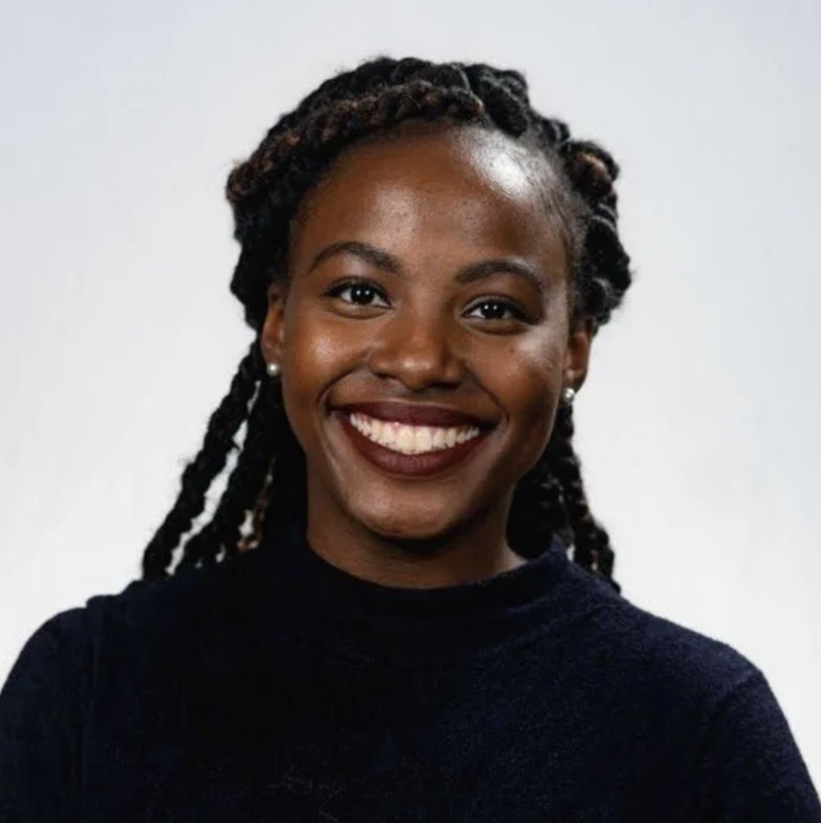 Portrait of a smiling Black woman with braids, wearing pearl earrings and a dark top, standing against a plain white background.