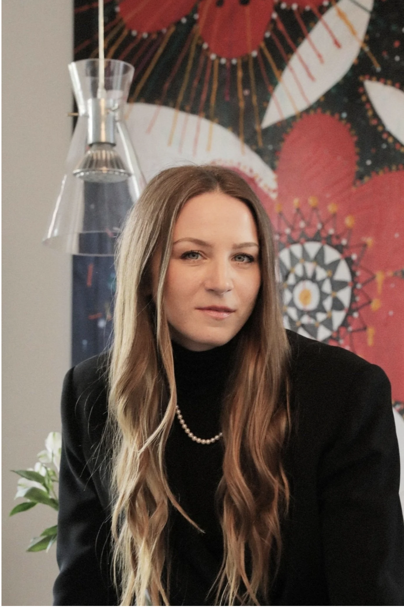 A woman with long, wavy brown hair wearing a black blazer and pearl necklace, sitting indoors with colorful artwork in the background and a modern glass lamp to her left.