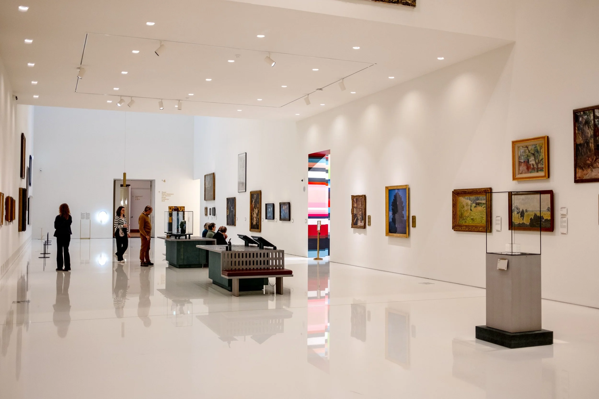 People viewing paintings and artwork in an art museum gallery with white walls and a glossy floor.