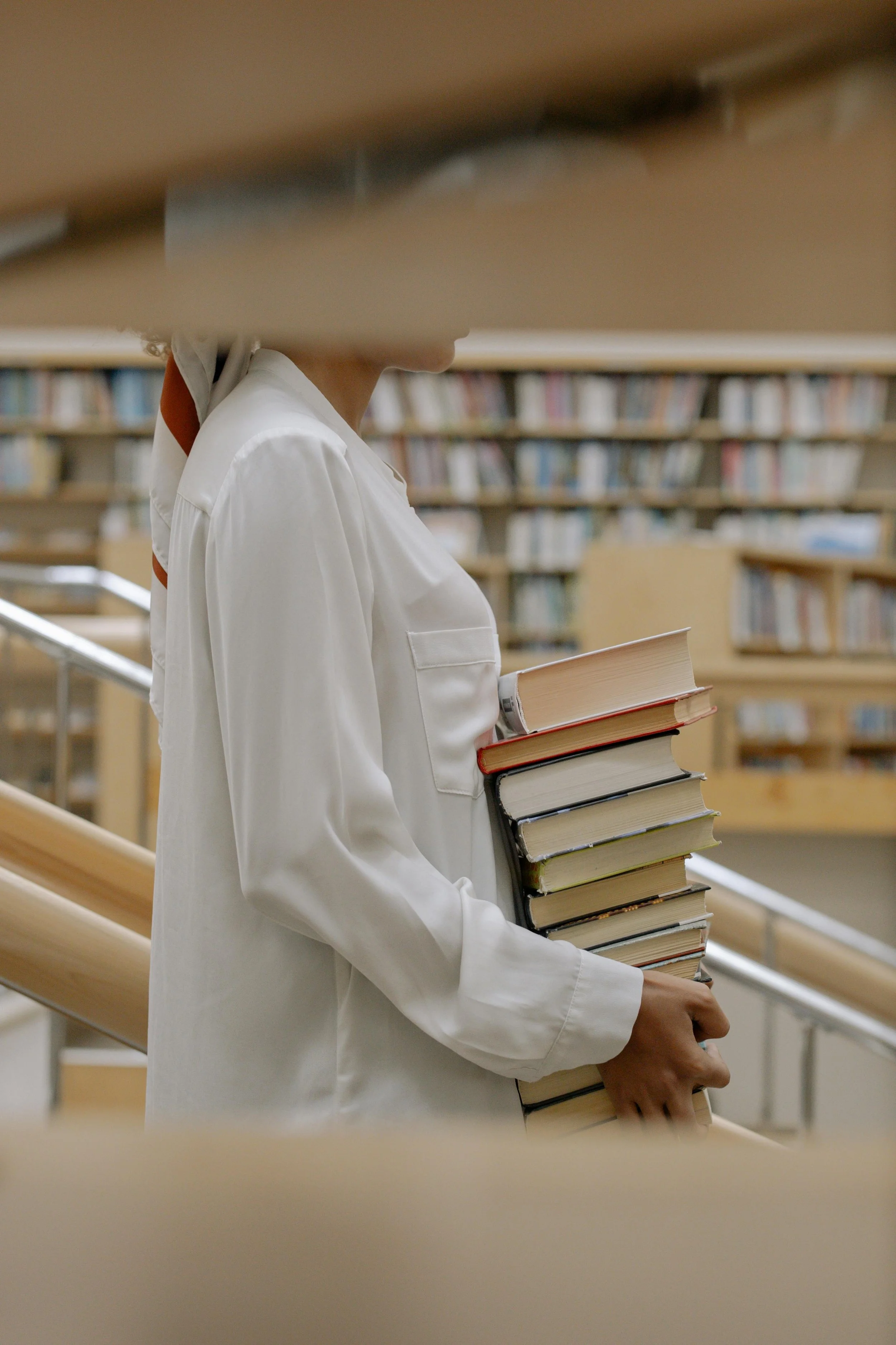 A person wearing a white jacket holding a stack of books in a library.