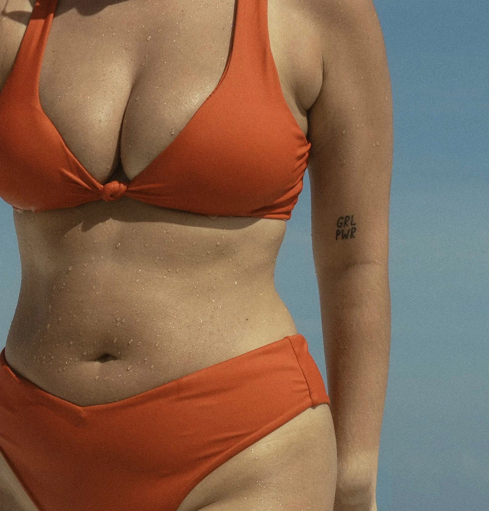 A woman wearing an orange bikini top tied in the front, with water droplets on her skin, standing against a blue background.