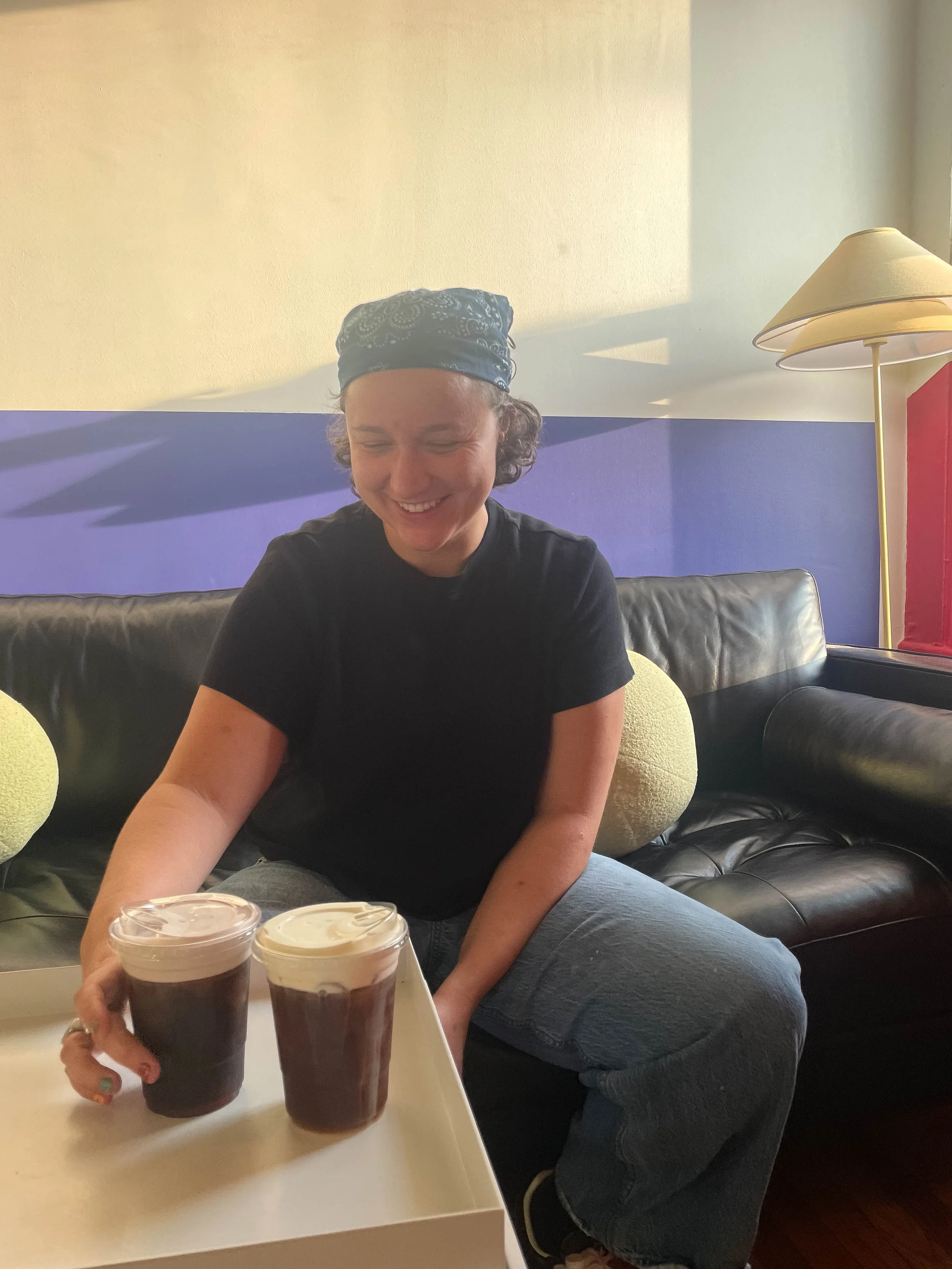 A person wearing a blue bandana smiling at a table with two cups of iced coffee, seated on a black leather couch in a room with a yellow and purple wall, a yellow floor lamp, and a red panel.