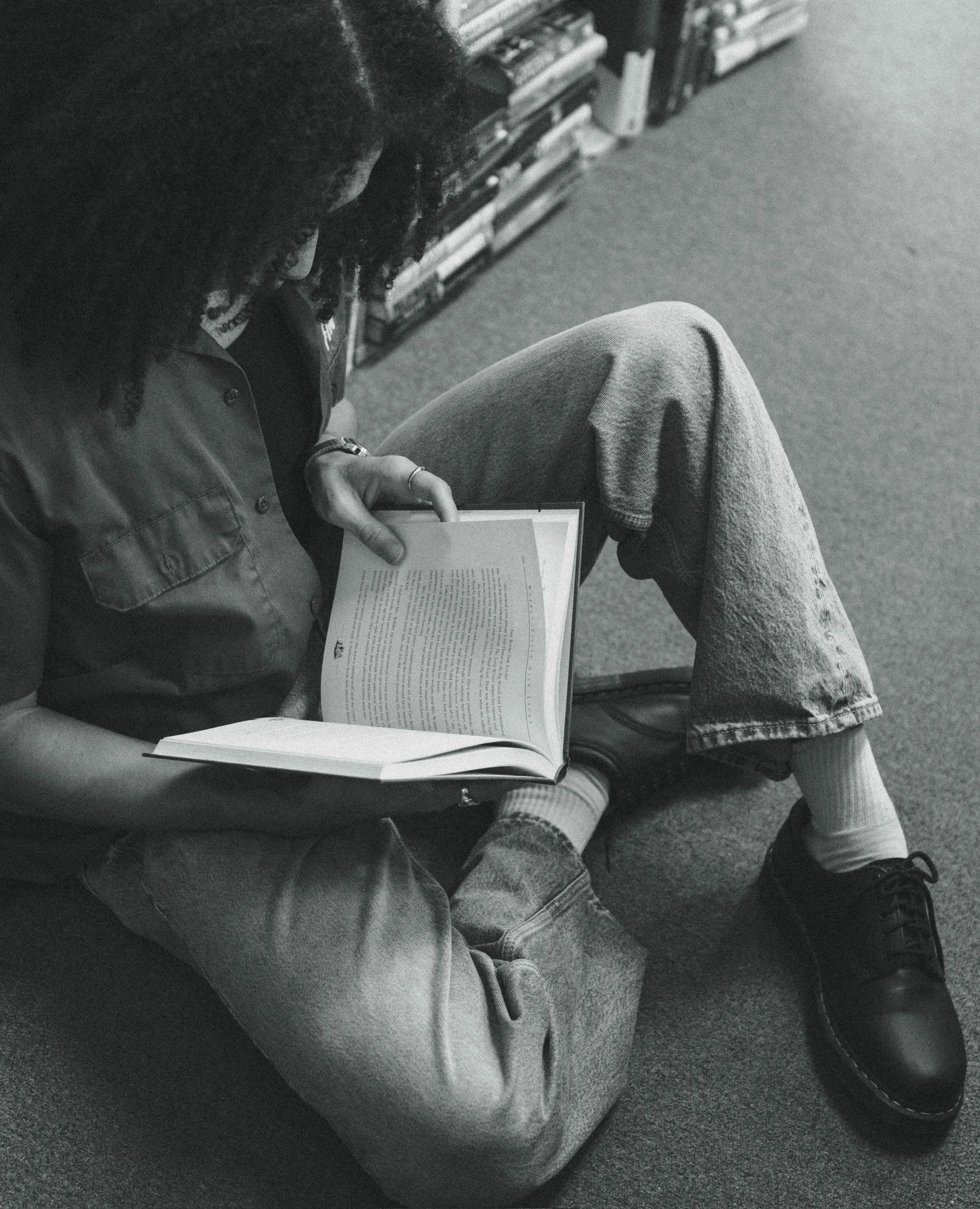 A person is sitting on the floor reading a book. They are wearing jeans, sneakers, and a jacket. There are bookshelves in the background.