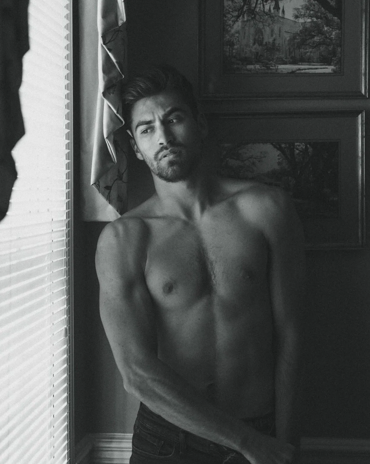 Black and white photo of a shirtless man with a beard, looking at the mirror with a serious expression, standing in front of a framed painting and a window with blinds.