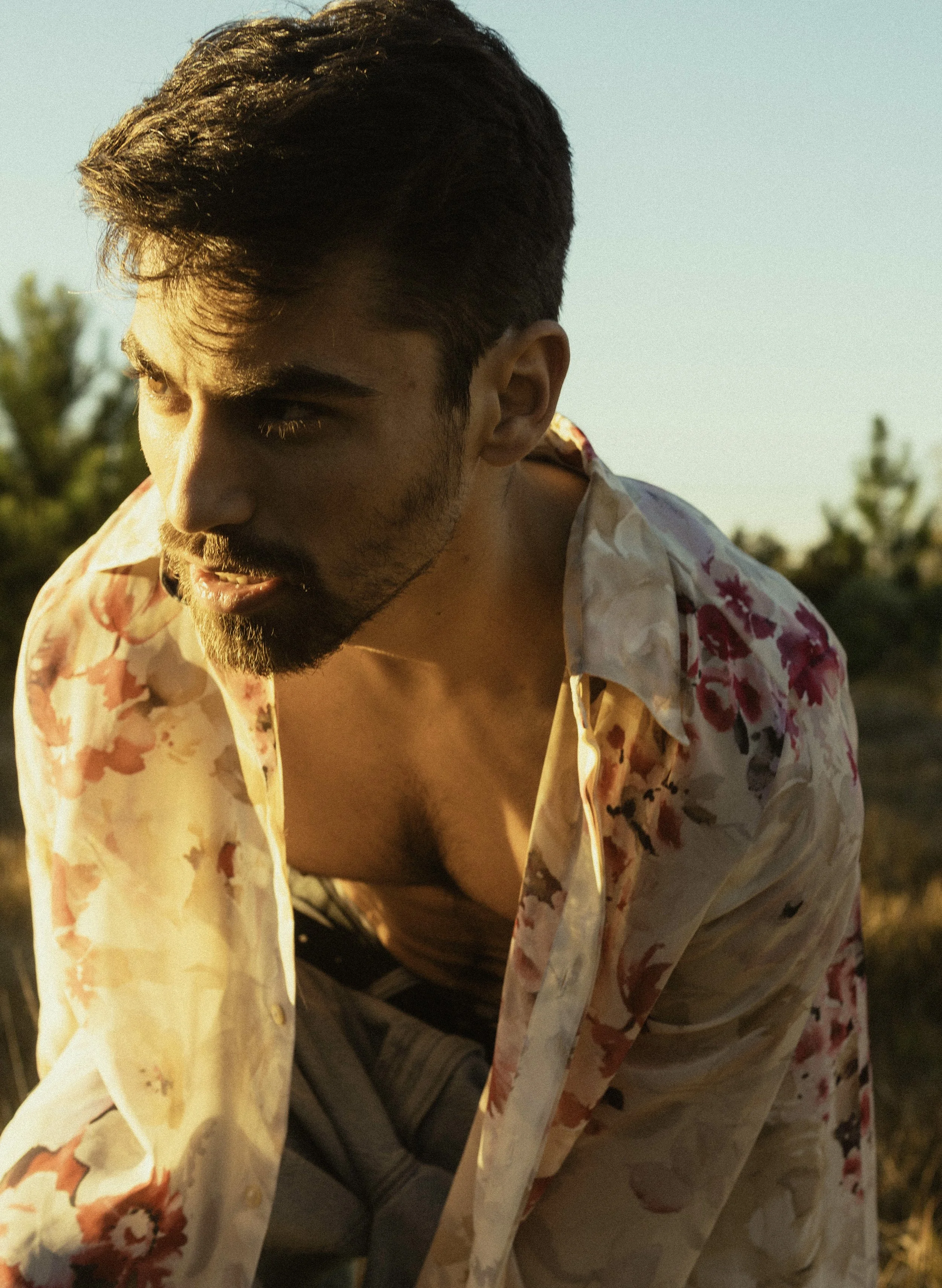A young man with dark hair and a beard wearing an open floral shirt, looking downwards during sunset or golden hour, with trees blurred in the background.
