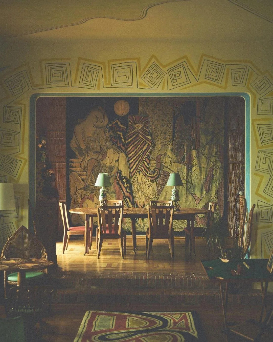 A cozy dining area with a wooden table, six chairs, two matching table lamps, and a large mural depicting abstract figures and designs on the wall. The room is decorated with hand-drawn spirals around the archway and a patterned rug on the floor.