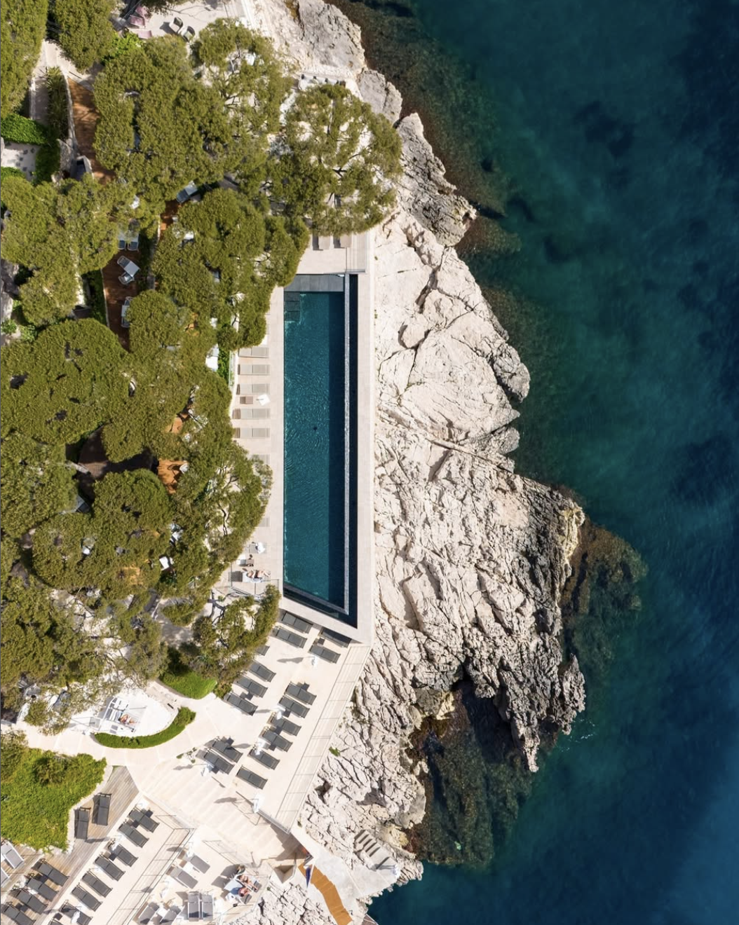 An aerial view of a rectangular swimming pool surrounded by lounge chairs, natural rocks, and lush green trees along a rocky coastline with clear blue water.